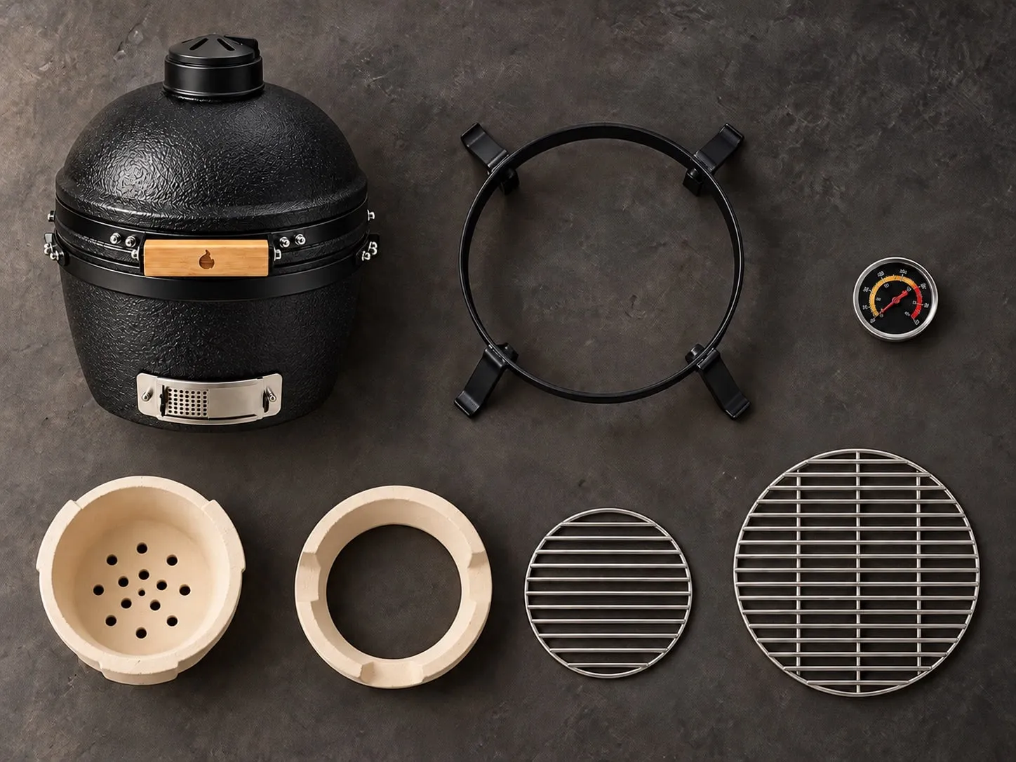 Flat-lay of everything in the 15″ box on dark concrete: the black kamado, stand, thermometer, ceramic fire box and fire ring, charcoal and cooking grates